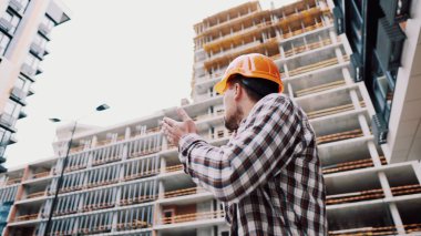 İnşaat alanında duran genç işçinin portresi. İnşaat alanında mühendis telefonda konuşuyor. Kask kontrolündeki yapıcı cep telefonuna göre planlandığı gibi.