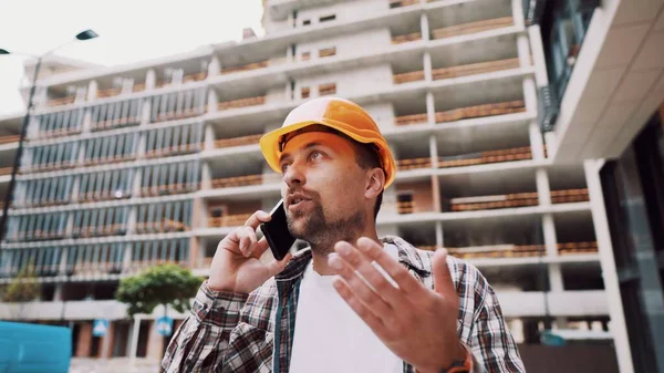 建設現場に立っている間に呼び出しを行う若い便利屋の肖像画 エンジニアが工事現場で電話で話してる 携帯電話による計画によるヘルメットコントロールのビルダー — ストック写真