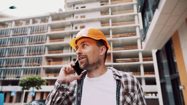 Caucasian male construction worker in orange hard hat and plaid shirt talking on phone at construction site. Architecture theme. Male profession. Foreman controls construction process by smartphone.