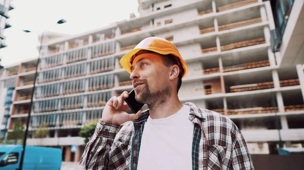 Caucasian male construction worker in orange hard hat and plaid shirt talking on phone at construction site. Architecture theme. Male profession. Foreman controls construction process by smartphone.