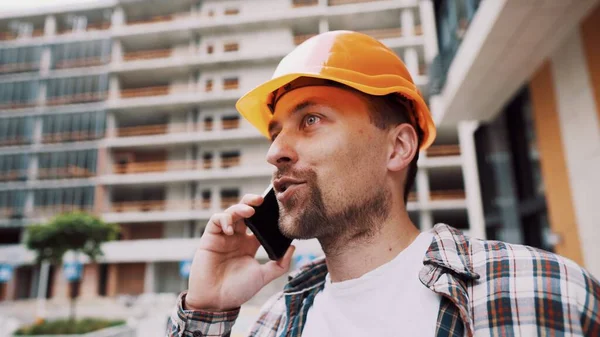 Caucasian male construction worker in orange hard hat and plaid shirt talking on phone at construction site. Architecture theme. Male profession. Foreman controls construction process by smartphone.