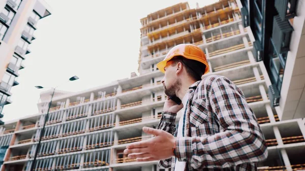 オレンジのハード帽子の白人男性建設労働者と建設現場で電話で話してシャツを果たした 建築のテーマ 男性専門職 フォアマンはスマートフォンで建設プロセスを制御します — ストック写真