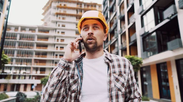 Caucasian male construction worker in orange hard hat and plaid shirt talking on phone at construction site. Architecture theme. Male profession. Foreman controls construction process by smartphone.