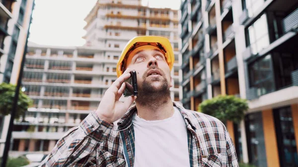 Portrait of young handyman making call while standing at construction area. Engineer talking on the phone on a construction site. Builder in helmet control according to plan by mobile phone.