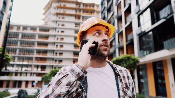 オレンジのハード帽子の白人男性建設労働者と建設現場で電話で話してシャツを果たした 建築のテーマ 男性専門職 フォアマンはスマートフォンで建設プロセスを制御します — ストック写真