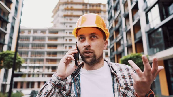 Caucasian male construction worker in orange hard hat and plaid shirt talking on phone at construction site. Architecture theme. Male profession. Foreman controls construction process by smartphone.