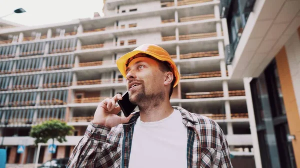 Portrait of young handyman making call while standing at construction area. Engineer talking on the phone on a construction site. Builder in helmet control according to plan by mobile phone.