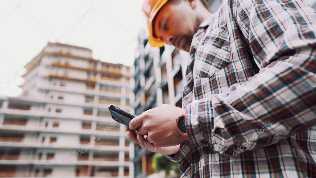 Young Engineer using smartphone on construction site. Architect using phone. Manager development building texting on smart phone. Builder makes notes in application during inspection and verification.