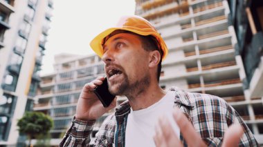 Angry builder, dissatisfied with deadline of work, swears at stress while talking to foreman on phone background of construction site. Wrathful architect, construction engineer talking on cell phone. clipart
