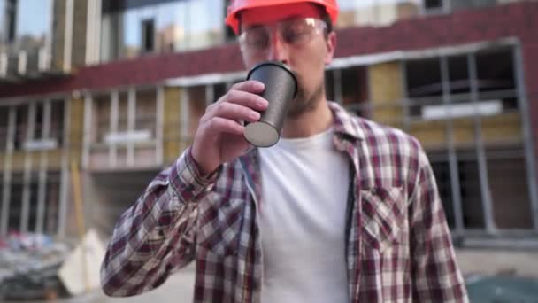 Jovem homem caucasiano construtor capataz cansado e bebe café no canteiro de obras de fundo. Engenheiro em equipamentos de proteção pausa para café. Inspetor de construção bebe bebida quente após o dia difícil — Vídeo de Stock