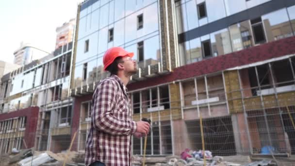 Junge kaukasische männliche Vorarbeiter Bauarbeiter müde und trinkt Kaffee auf Hintergrundbaustelle. Ingenieur in Schutzausrüstung Kaffeepause. Bauinspektor trinkt nach hartem Tag Heißgetränk — Stockvideo