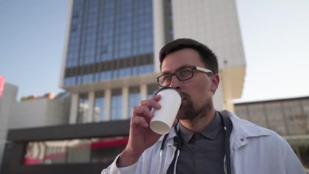 하루 종일 커피를 마시며 휴식을 취하는 전문 의료인. 일반 개업 의사는 진료소의 뒤를 걷는 동안 커피를 마십니다. 실험실 코트를 입은 의사가 따뜻 한 음료를 마신다 — 비디오