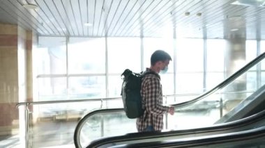 Beyaz maskeli adam boş bir havaalanı terminalinde yürüyen merdivene tırmanıyor. Salgın Corona Covid 19 virüsü sırasında seyahat et. Güvenli mesafede kalın. Maskeli bir erkek uçakla gitmek için kapıya gidiyor.