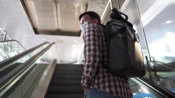 旅行保険の概念。空港ターミナルでエスカレーターにバックパックを運ぶ白人観光客の男性の顔マスク。旅行中のCOVID-19コロナウイルスパンデミックの予防。安全のために。新常態 — ストック動画