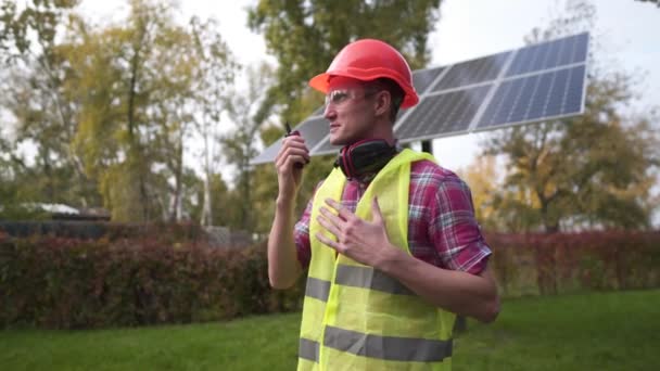 Trabalhador masculino caucasiano em um capacete vermelho, colete verde e equipamento de proteção serve um pequeno painel solar. O engenheiro fala e dá comandos sobre o rádio na central solar — Vídeo de Stock