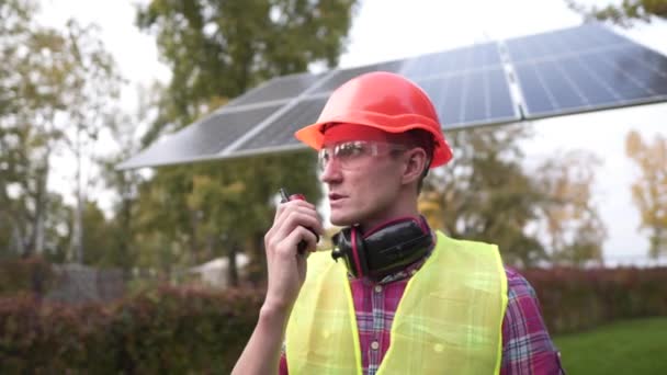 Καυκάσιος εργάτης με κόκκινο κράνος, πράσινο γιλέκο και προστατευτικό εξοπλισμό εξυπηρετεί ένα μικρό ηλιακό πάνελ. Ο μηχανικός μιλάει και δίνει εντολές από το ραδιόφωνο στο σταθμό ηλιακής ενέργειας — Αρχείο Βίντεο
