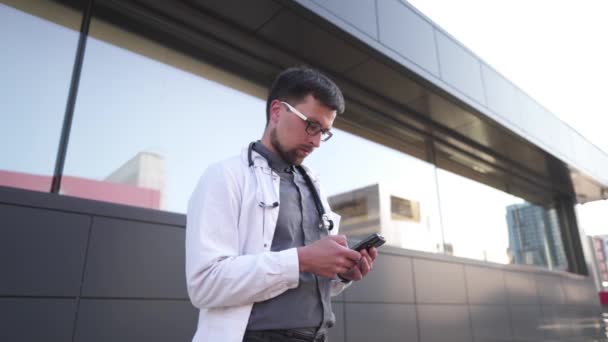 A szemüveges doktor üzenetet kap az okostelefonon. Fiatal háziorvos telefonál szünetben. Az orvos mobiltelefont használ a szabadban. Doki a kórház előtt áll mobiltelefonnal. — Stock videók