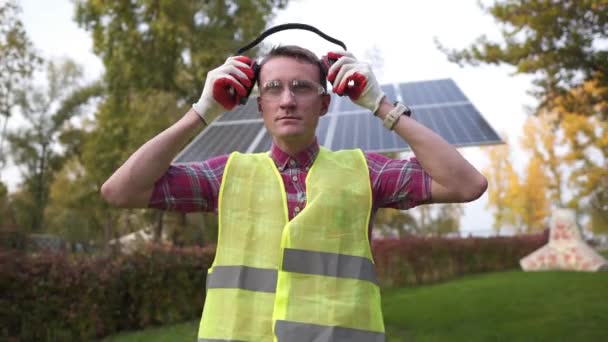 Um engenheiro com fones de ouvido trabalha na estação solar. Trabalhador coloca fones de ouvido para proteção sonora perto do painel solar. Cuidar da saúde, segurança no trabalho. Tampões auriculares. Som muito alto, deficiência auditiva — Vídeo de Stock