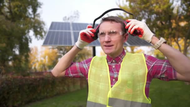 Técnico ou engenheiro com fones de ouvido de proteção em pé perto do painel solar. Homem usando proteção auditiva de equipamentos de segurança. Trabalhador usando defensores do ouvido de cancelamento de ruído ou abafadores de ouvido — Vídeo de Stock