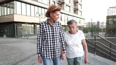 Aile, nesil ve insan kavramı. Mutlu gülümseyen olgun bir kız ve yaşlı bir anne şehrin modern evinin avlusunda yürüyor. Olgun anne ve yetişkin kız cadde boyunca kucaklaşarak yürür.