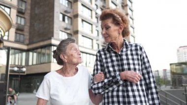 Aile, nesil ve insan kavramı. Mutlu gülümseyen olgun bir kız ve yaşlı bir anne şehrin modern evinin avlusunda yürüyor. Olgun anne ve yetişkin kız cadde boyunca kucaklaşarak yürür.