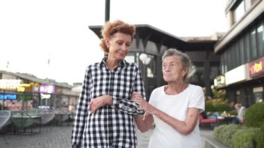 Aile, nesil ve insan kavramı. Mutlu gülümseyen olgun bir kız ve yaşlı bir anne şehrin modern evinin avlusunda yürüyor. Olgun anne ve yetişkin kız cadde boyunca kucaklaşarak yürür.