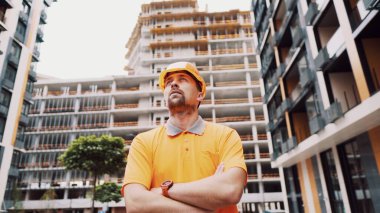 Güvenlik kaskı ve turuncu yansıtıcı üniformayla kolları çaprazlanmış, inşaat alanında gururlu pozlar veren Kafkasyalı genç bir inşaatçı. İnşaat alanında poz veren gururlu bir işçi. İşçi portresi.