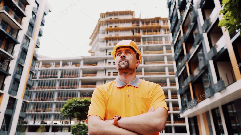 Young caucasian constructor with arms crossed in safety helmet and orange reflective uniform posing with proud expression at construction site. Proud worker posing on building site. Worker portrait.