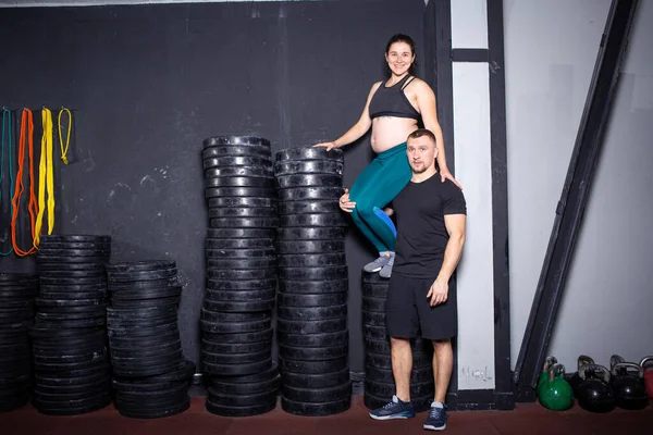 Workouts and exercises for pregnant women in the gym. Caucasian athletes bodybuilders, couple husband and wife enjoy pregnancy and posing after training near barbell and dumbbell equipment.