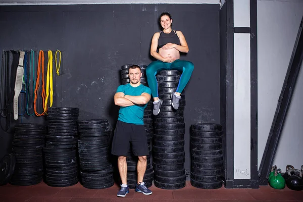 Workouts and exercises for pregnant women in the gym. Caucasian athletes bodybuilders, couple husband and wife enjoy pregnancy and posing after training near barbell and dumbbell equipment.