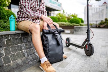 Beyaz bir genç, şehir parkında elektrikli bir scooter 'ın yanındaki bankta otururken siyah bir sırt çantası açıyor. Kurye, teslimat hizmeti konsepti. Sırt çantalı bir erkek açık bir sırt çantasına bakar..