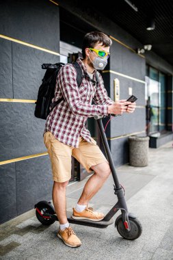 Koruyucu yüz maskesi takan erkek kurye, elektrikli scooterla teslimat adresine ulaştı. Çevre dostu taşımacılıkta hedef 19 sırasında teslim edildi. Büyük şehirde kirlilik. Başbakan 2.5 maskesi.