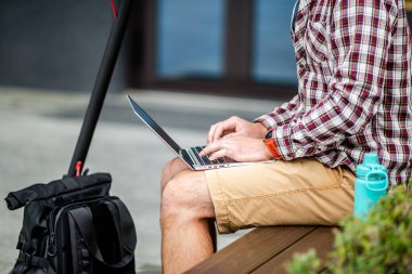 Merkezdeki ahşap bankta otururken dizüstü bilgisayarın klavyesinde daktilo yazarken yakın çekim. Erkek serbest çalışan dizüstü bilgisayar ve elektrikli scooter kullanıyor. Mesaj, fikir. Çalışma alanı herhangi bir eşya.