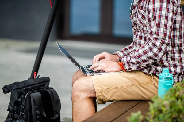 Merkezdeki ahşap bankta otururken dizüstü bilgisayarın klavyesinde daktilo yazarken yakın çekim. Erkek serbest çalışan dizüstü bilgisayar ve elektrikli scooter kullanıyor. Mesaj, fikir. Çalışma alanı herhangi bir eşya.