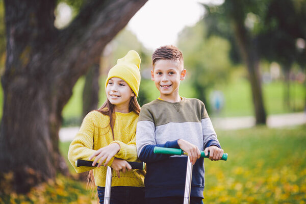 Two children caucasian boy and girl twins posing with scooters in autumn park. Happy kids riding kick scooters in public park. Kids sport. Outdoor activities brother sister teens. Happy childhood