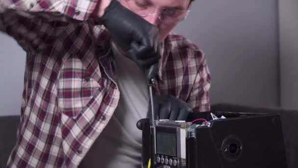 Um jovem está tentando consertar seus próprios aparelhos de cozinha em casa, uma cafeteira. Homem caucasiano repara uma máquina de café com uma chave de fenda vestida com uma camisa xadrez e óculos e luvas de segurança — Vídeo de Stock