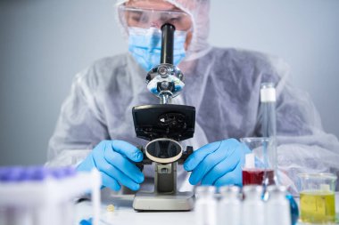 Bilim adamı mikroskopla numune inceliyor, bilim laboratuarında tıbbi araştırma yapıyor. Koruyucu giysiler içindeki erkek laboratuvar asistanı laboratuvarda mikroskopla yeni kimyasal maddeler üzerinde çalışıyor..