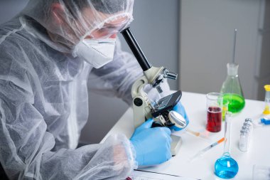 Bilim adamı mikroskopla numune inceliyor, bilim laboratuarında tıbbi araştırma yapıyor. Koruyucu giysiler içindeki erkek laboratuvar asistanı laboratuvarda mikroskopla yeni kimyasal maddeler üzerinde çalışıyor..
