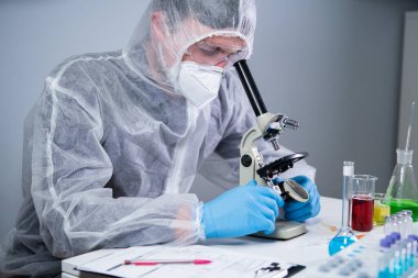 Bilim adamı mikroskopla numune inceliyor, bilim laboratuarında tıbbi araştırma yapıyor. Koruyucu giysiler içindeki erkek laboratuvar asistanı laboratuvarda mikroskopla yeni kimyasal maddeler üzerinde çalışıyor..