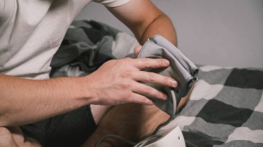 Beyaz bir genç yatak odasındaki kan basıncını ölçüyor. Ev kıyafetleri içinde yatakta oturuyor. Erkek, kan basıncını ölçmek için tonometre kullanıyor. Genç yaşta yüksek tansiyon.