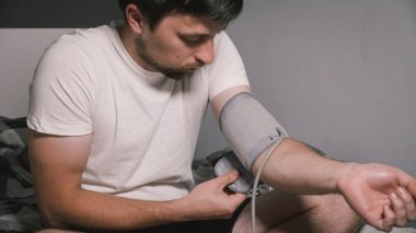 Beyaz bir genç yatak odasındaki kan basıncını ölçüyor. Ev kıyafetleri içinde yatakta oturuyor. Erkek, kan basıncını ölçmek için tonometre kullanıyor. Genç yaşta yüksek tansiyon.
