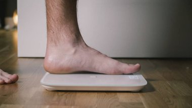Adam ağırlığı kontrol etmek için dijital cam teraziye basıyor. Erkek, ev ortamında zemin tartması yapıyor, ayak manzarasını kapatıyor. Elektronik terazide erkek ayağı. Spor konsepti. Zayıflama ve sağlıklı yaşam tarzı.