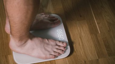İnce adamın çıplak bacakları beyaz banyo terazisine basar. Erkek evde antrenmandan sonra kilosunu kontrol eder. Adam ağırlık ve yağ oranını dijital ağırlık ve sağlıklı yaşam tarzına göre kaydediyor..