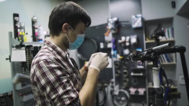 Mecánica de reparación de bicicletas en taller con máscara médica covid 19. Hombre bicicleta de reparación en cuarentena pandemia coronavirus en máscara facial protectora. El hombre examina y arregla el ciclo. Concepto tienda deporte — Vídeo de stock