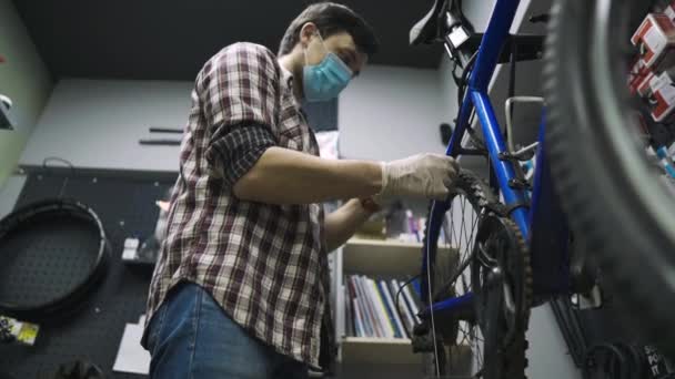 Mechanická oprava jízdního kola v dílně s kovidem lékařské masky19. Muž oprava kolo na karanténní coronavirus pandemie v ochranné masce obličeje. Člověk zkoumá a opravuje cyklus. Koncept sportovního obchodu — Stock video