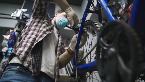 El mecánico de bicicletas repara y mantiene la bicicleta del cliente de acuerdo con los estándares de cuarentena durante la pandemia de coronavirus, usa máscara médica y guantes. Transporte individual durante el bloqueo covid 19 — Vídeos de Stock