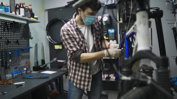 Mécanicien réparateur vélo en atelier portant un masque médical covide 19. Homme réparation vélo sur la pandémie de coronavirus en quarantaine dans un masque protecteur. L'homme examine et fixe le cycle. Concept de magasin de sport — Video