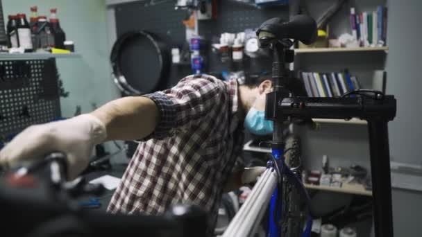 Mechanik naprawy rowerów w warsztacie w masce medycznej covid 19. Męski rower naprawczy na pandemii koronawirusowej w masce ochronnej. Człowiek bada i naprawia cykl. Koncepcja sklepu sportowego — Wideo stockowe