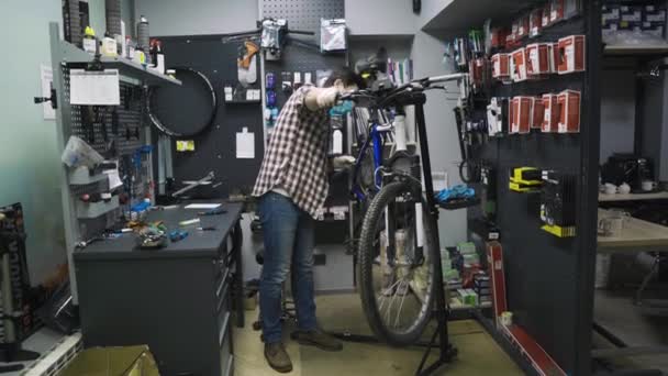 Mécanicien réparateur vélo en atelier portant un masque médical covide 19. Homme réparation vélo sur la pandémie de coronavirus en quarantaine dans un masque protecteur. L'homme examine et fixe le cycle. Concept de magasin de sport — Video