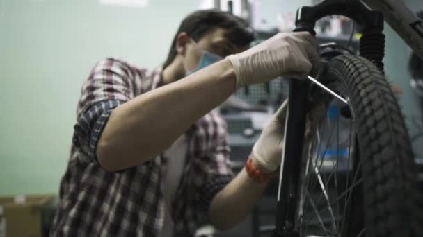 Сервисный инженер проводит диагностику и ремонт велосипеда в профессиональной мастерской, надев защитную маску и защитные перчатки от клиентского ковыда 19. Тема обслуживания во время пандемии коронавируса — стоковое видео